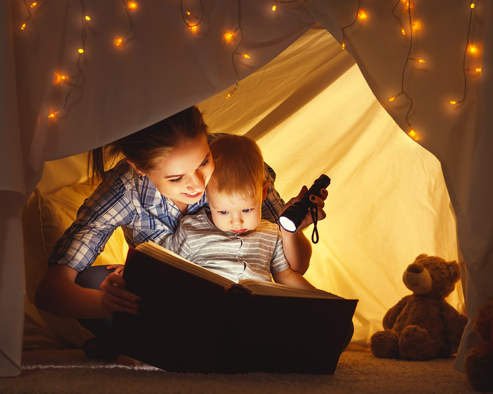 A mom and her toddler son sitting in a blanket fort with twinkle lights while she reads to him from a book with a flashlight