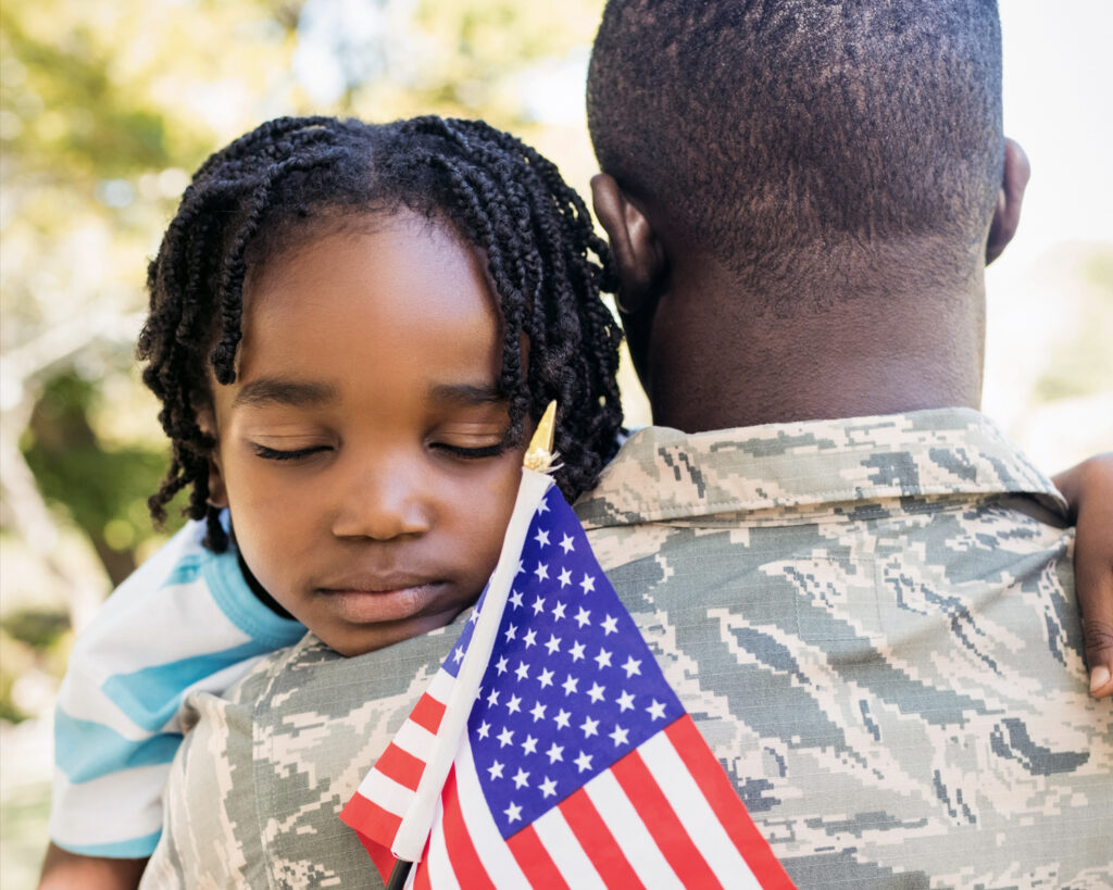 a Veteran holding his son who is sleeping on his shoulder holding an american flag